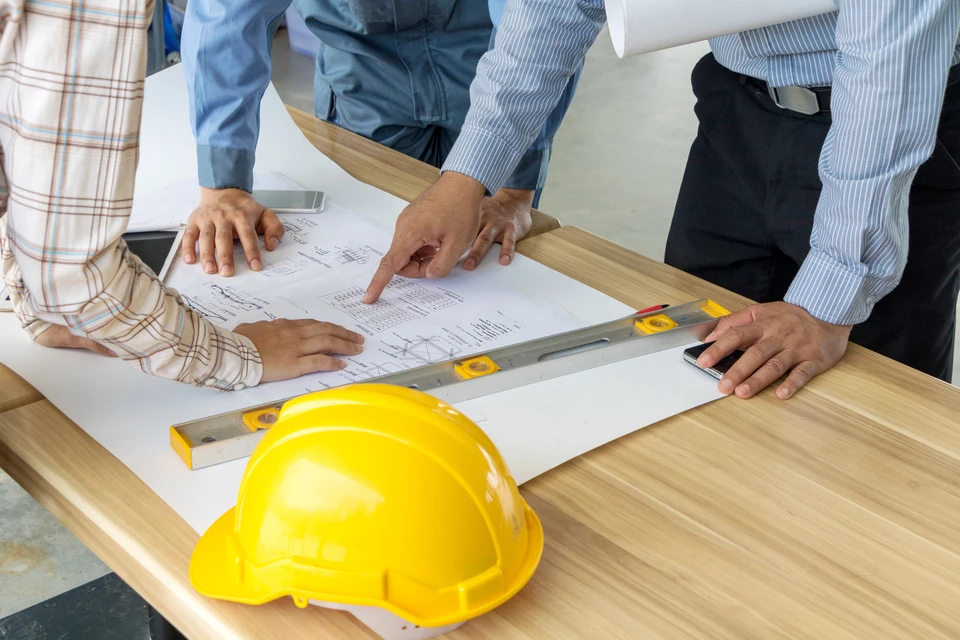 Réunion de travail en intérieur avec trois professionnels analysant des plans techniques sur une table en bois